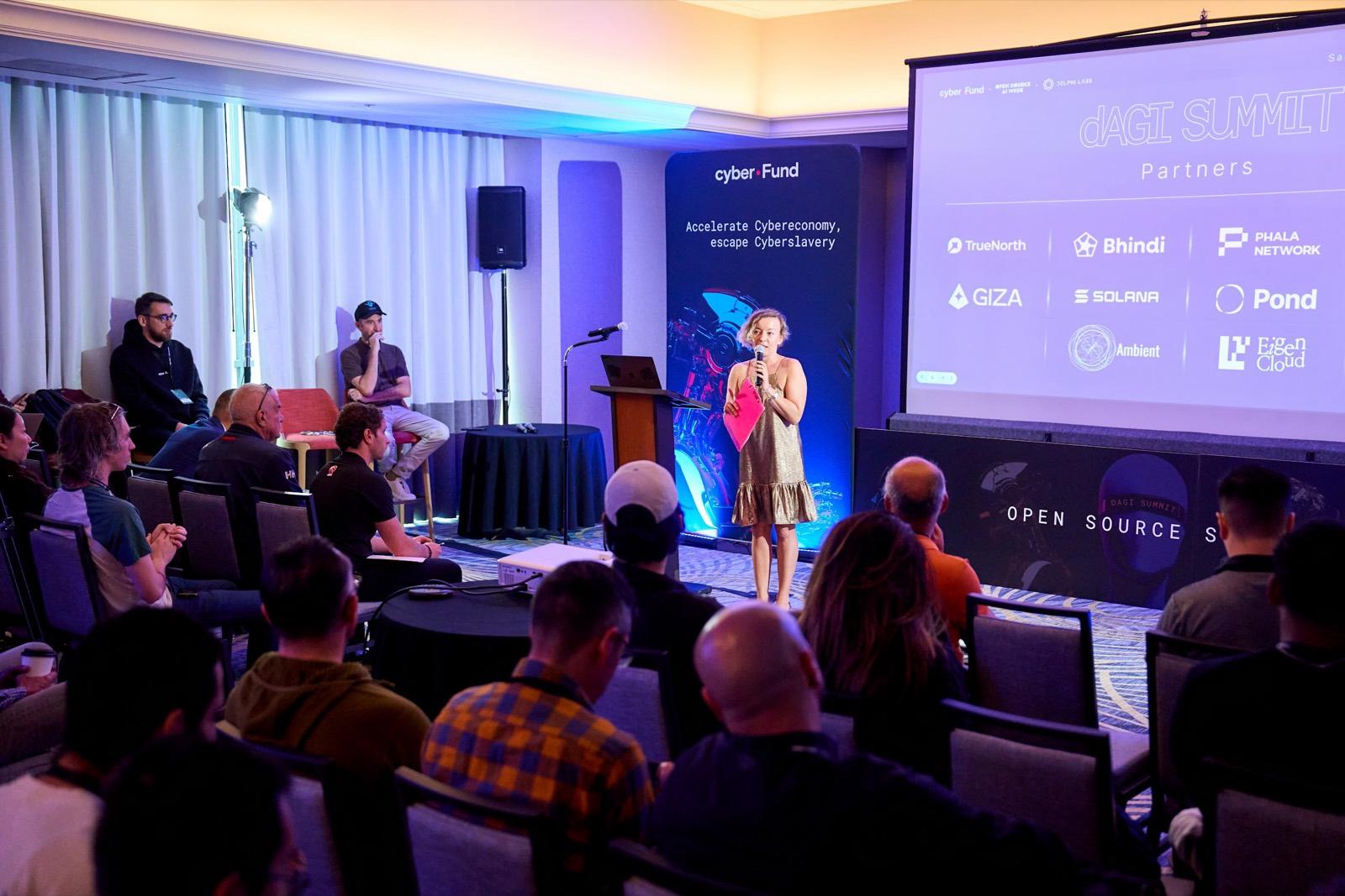 A speaker presenting on stage at the dAGI Summit in front of a seated audience and partner logo wall